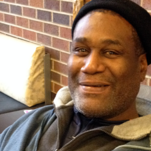 An African American man with a black cap on, sitting and smiling at the camera